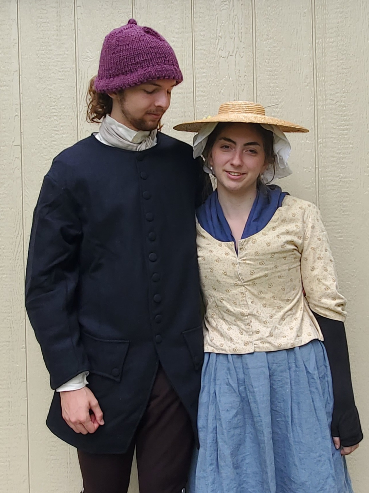 Young Couple of 18th Century Reenactors. The man is wearing a mid 18th Century Sleeved Weskit made with Navy Blue wool and a wine color hand knit Monmouth Cap. The young woman is wearing a light blue linen petticoat, a yellow cotton jacket, a white linen ruffled bonnet topped with a straw hat, a navy blue linen kerchief and black silk fingerless mittens.